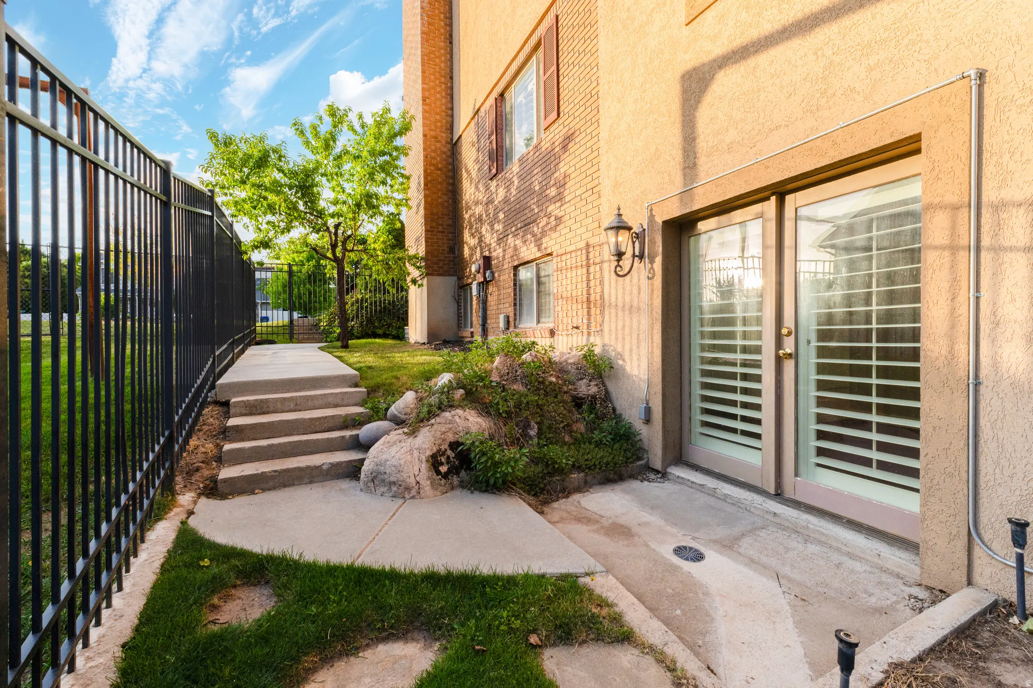 Exterior featuring a stucco finish, a black metal fence, concrete pathway with steps, and a grass area with mature trees