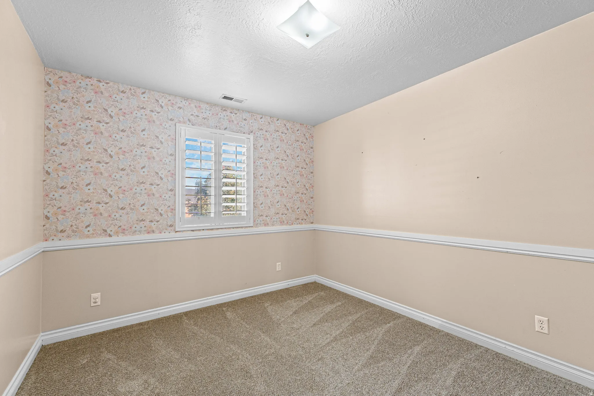 Room featuring a window with plantation shutters, patterned wallpaper, chair rail molding, and textured carpeting