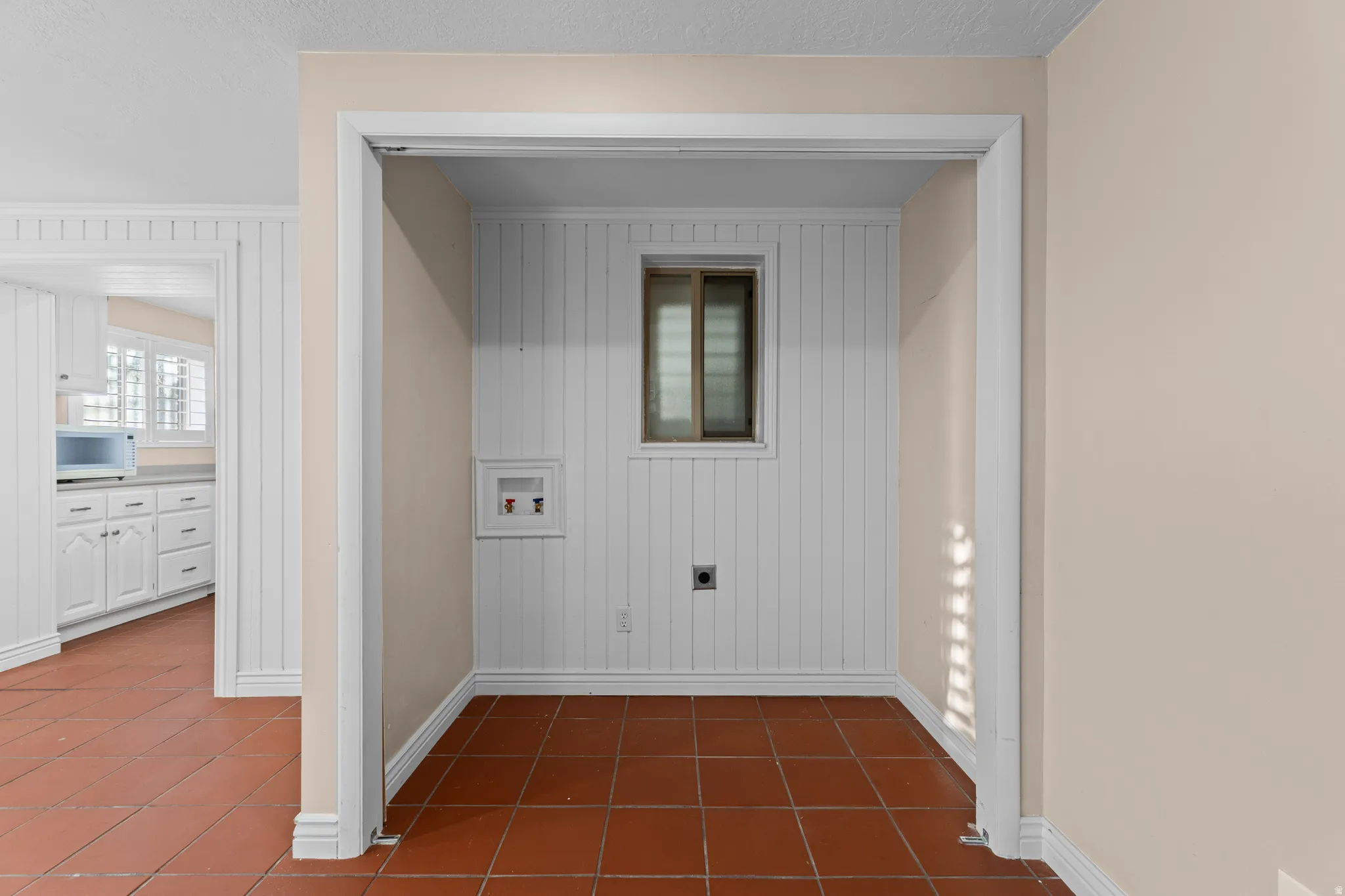 Dedicated utility space featuring terra cotta tile flooring, beadboard wall paneling, a single window, and visible plumbing connections