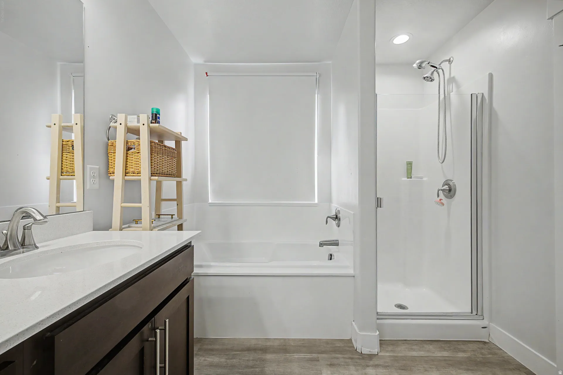 Bathroom featuring a dark wood vanity with a white countertop and undermount sink, a white bathtub with a window above, a separate shower enclosure with a glass door, and wood-finish flooring