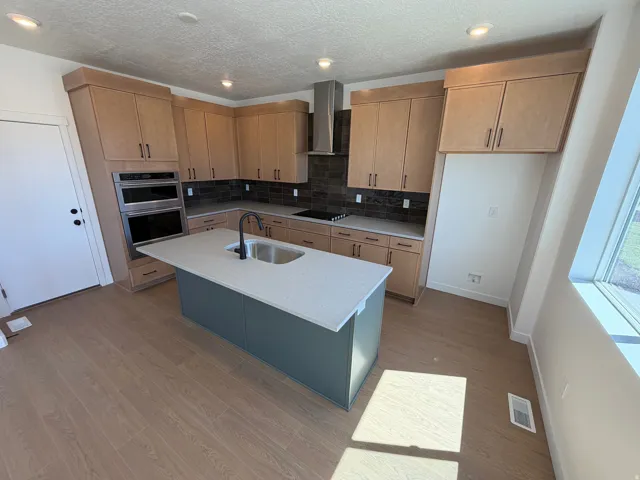 Modern kitchen featuring light wood-finish cabinetry, a central island with a sink, stainless steel double ovens, and a dark tile backsplash