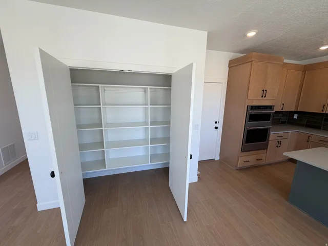 Custom pantry with extensive built-in shelving, wood-finish flooring, light wood cabinetry, stainless steel double wall ovens, and a kitchen island with a light-colored countertop