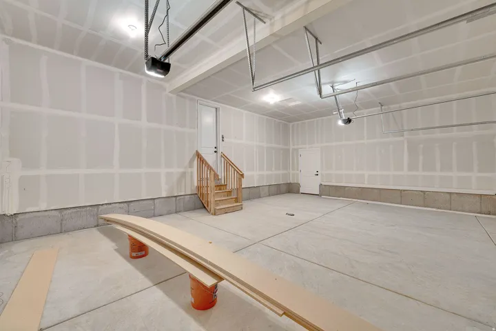 Spacious garage featuring a concrete floor, exposed drywall, overhead lighting, and an internal access door with wooden stairs and railing