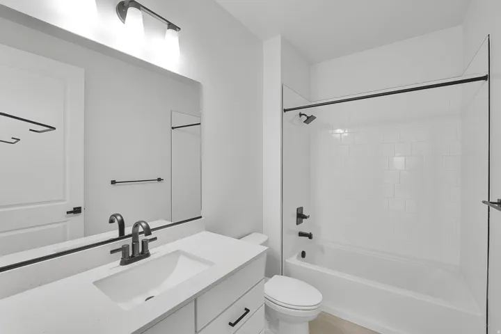 Bathroom featuring a single vanity with an integrated sink and light-toned countertop, matte black fixtures, a wall-mounted mirror, and a shower-over-tub combination with white subway tile surround and a glass enclosure