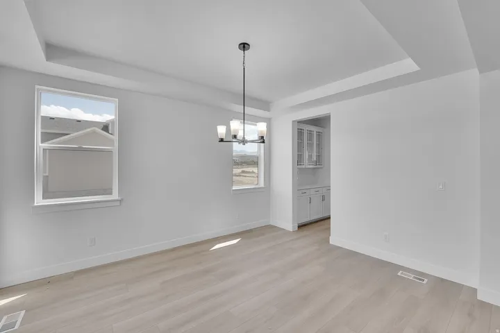 Spacious room featuring tray ceiling, contemporary chandelier, two windows, light wood-finish flooring, and an adjacent area with built-in cabinetry