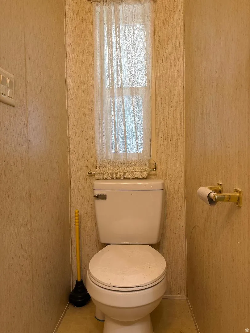 1/2 BathroomWhite commode with a window above, featuring light-colored wall paneling and a brass-tone toilet paper holder