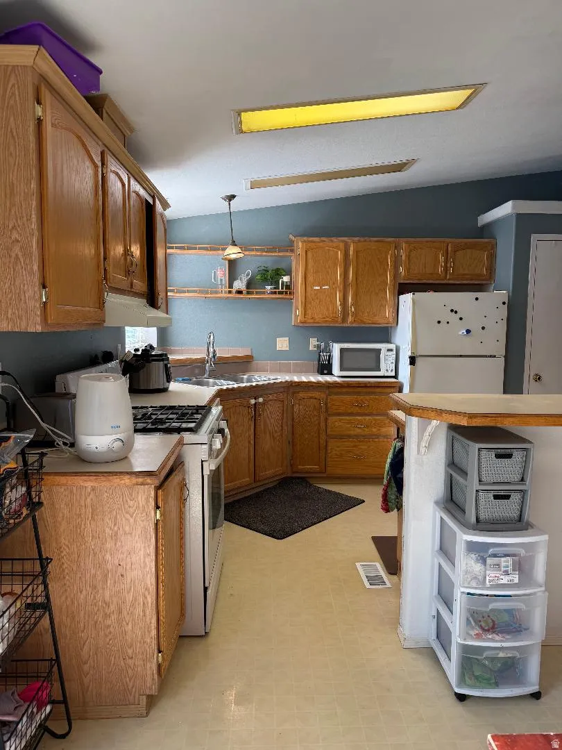 Kitchen featuring wood-finish cabinetry, a built-in oven range, a double basin sink, light-toned flooring, and wall-mounted shelving