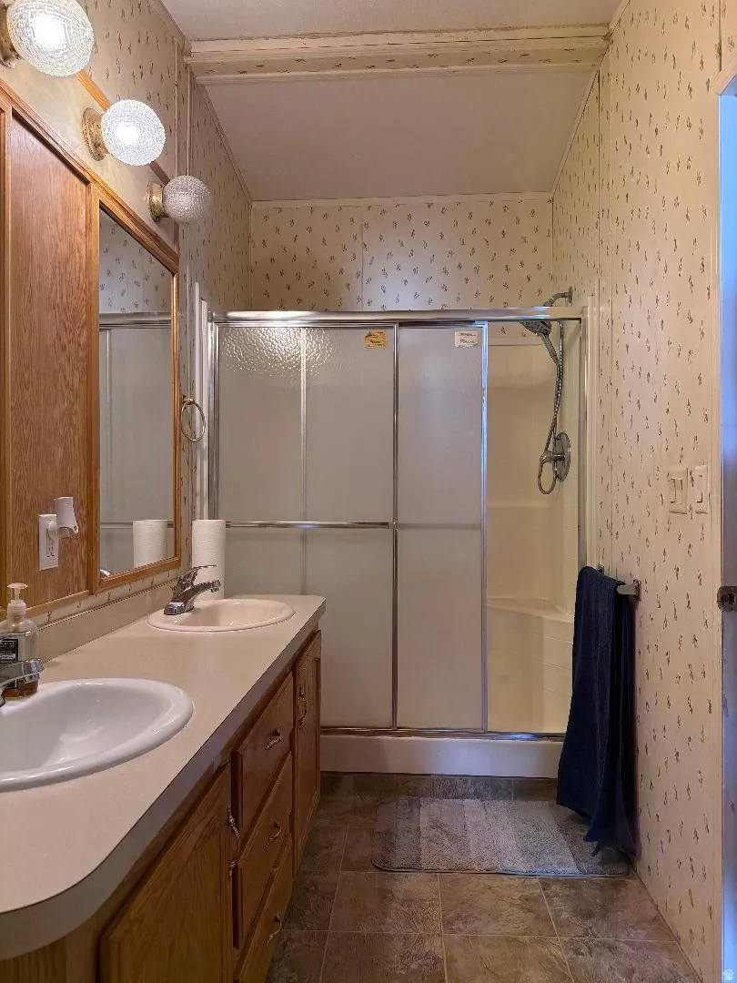Master Bathroom featuring a dual-sink vanity with wood cabinetry, a shower with sliding frosted glass doors, and patterned wallpaper