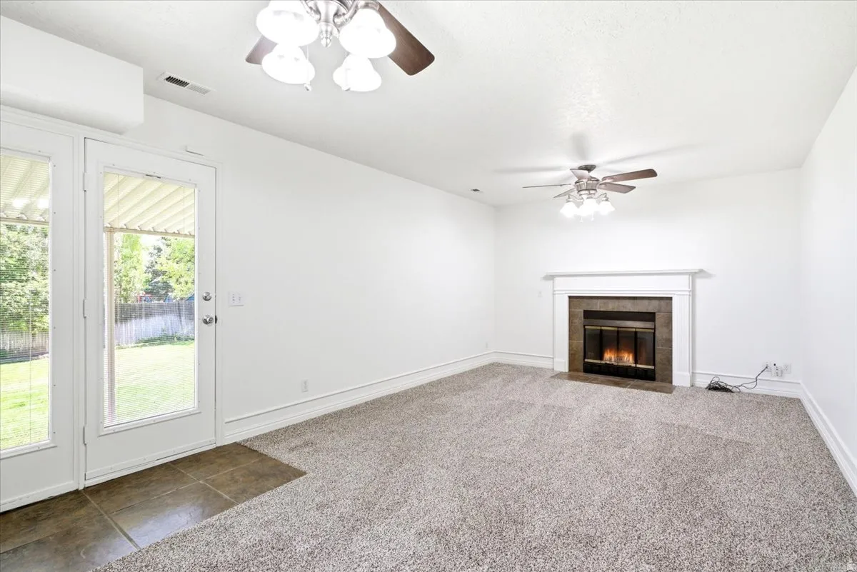 Spacious room featuring a fireplace with a white mantel, ceiling fans, light-toned carpet flooring, and a glass-paneled double door leading to an outdoor area