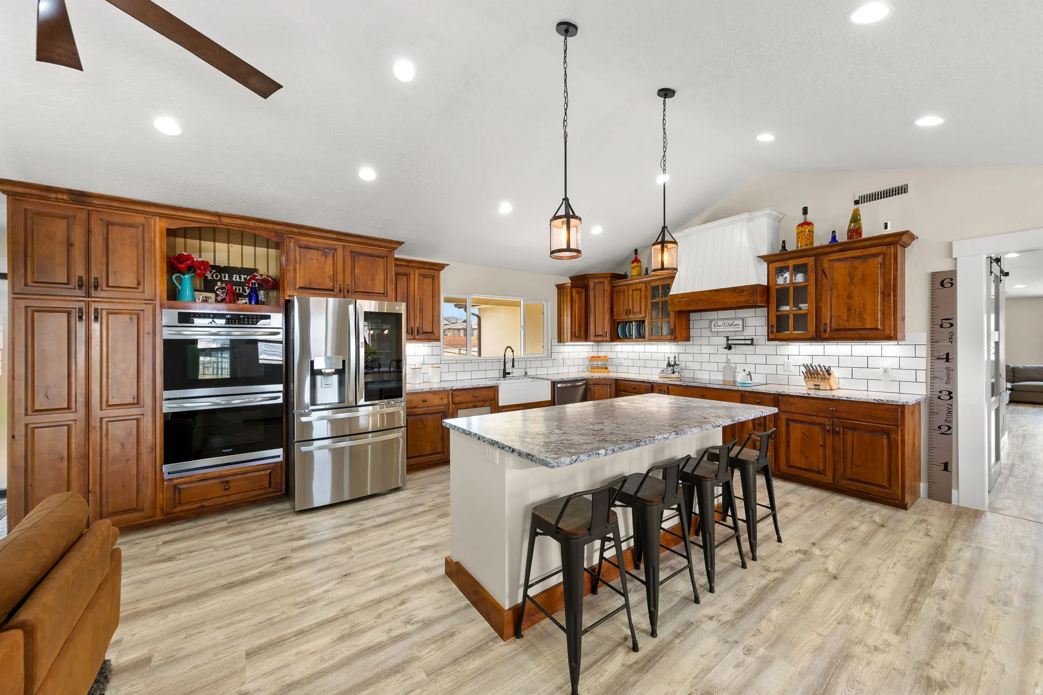 Spacious kitchen featuring an expansive center island with seating, wood-finish flooring, stainless steel appliances, white subway tile backsplash, and rich wood cabinetry