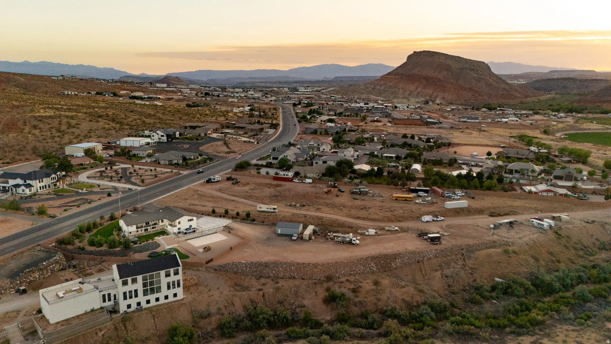 Panoramic aerial perspective featuring a modern white-exterior structure with extensive windows, a paved driveway, and surrounding arid landscape