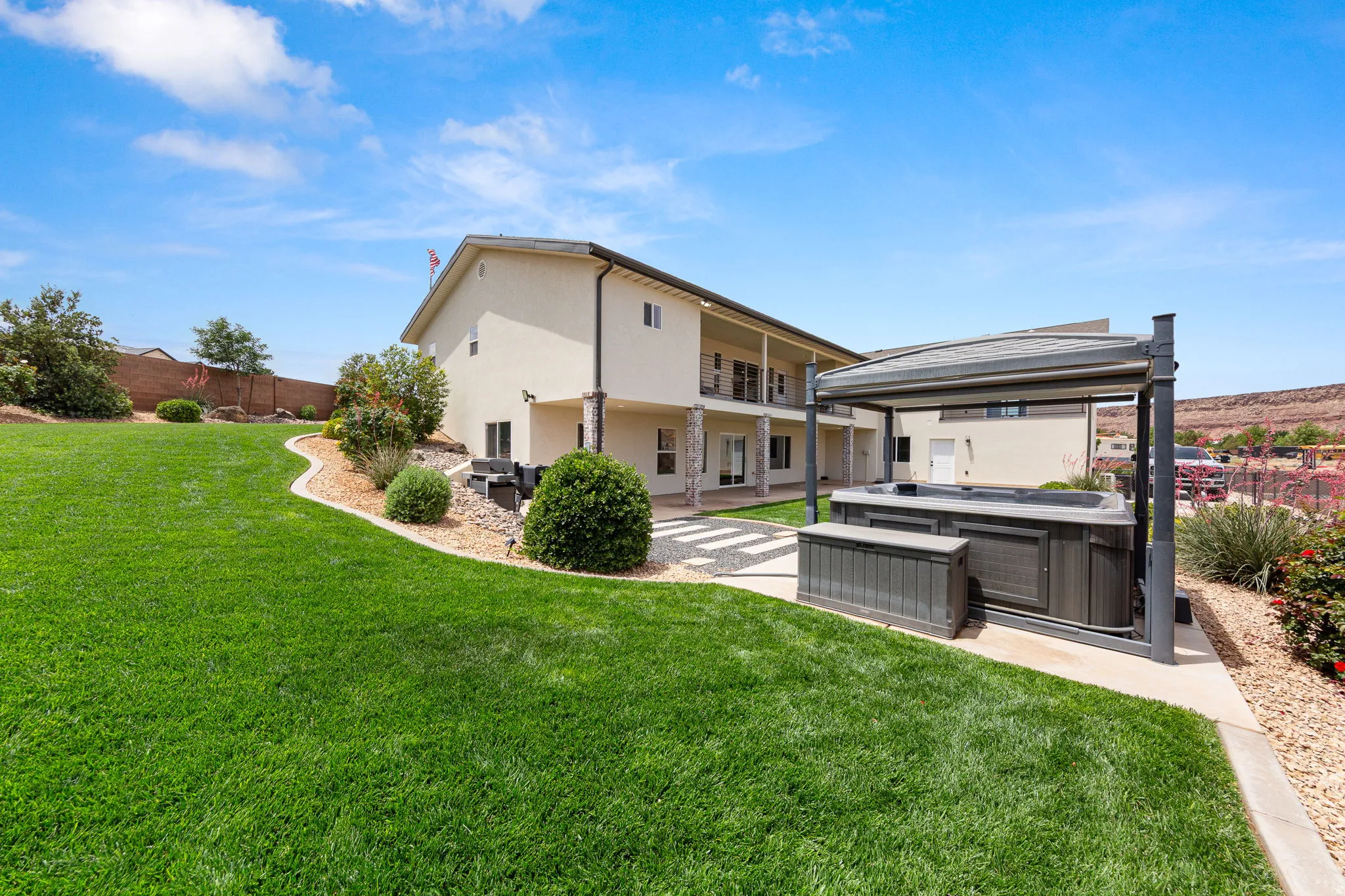 Expansive backyard featuring a lush green lawn, a covered hot tub area with a privacy screen, and a stone-paver pathway leading to the lower level patio