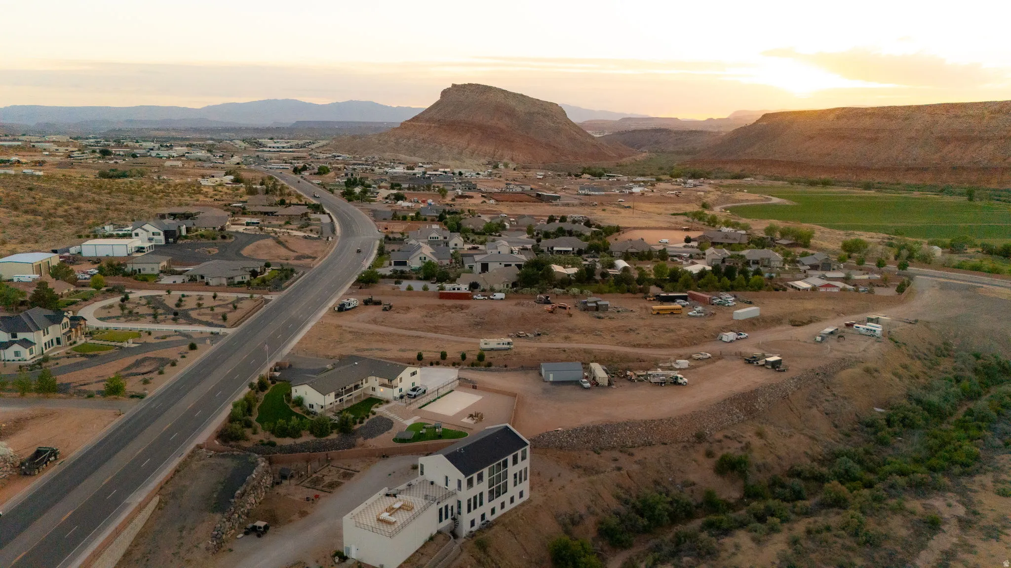 Expansive high desert landscape featuring a residential community, a winding paved road, and distant mountain formations