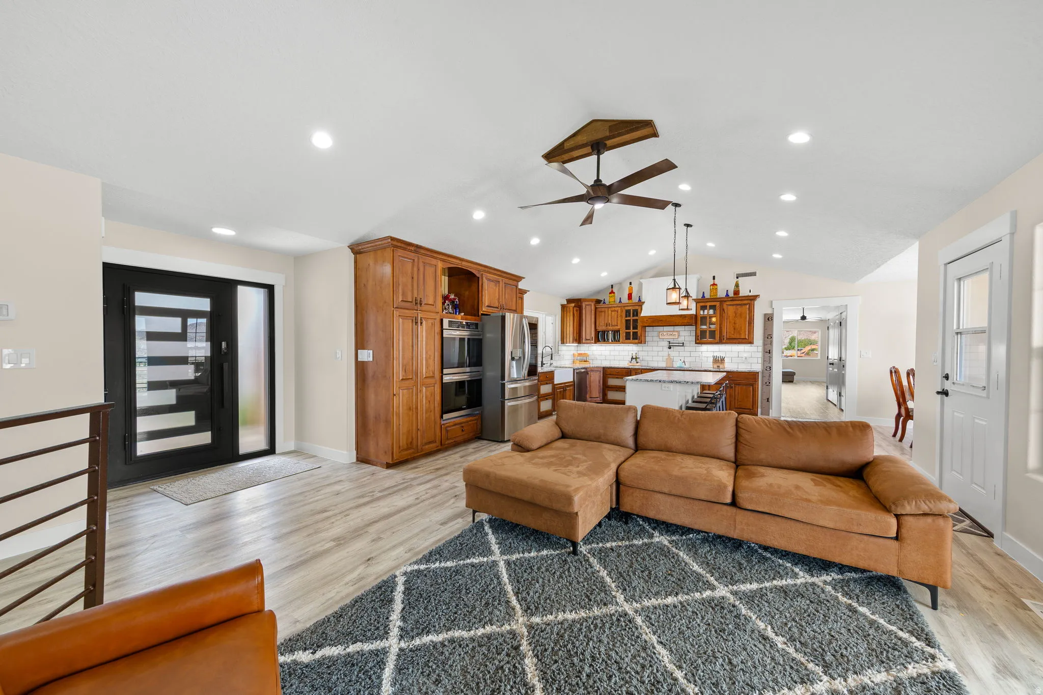 Open-concept living area featuring wood-finish flooring, a contemporary ceiling fan, recessed lighting, a modern entry door with frosted glass, and a kitchen with warm wood cabinetry and an island with seating