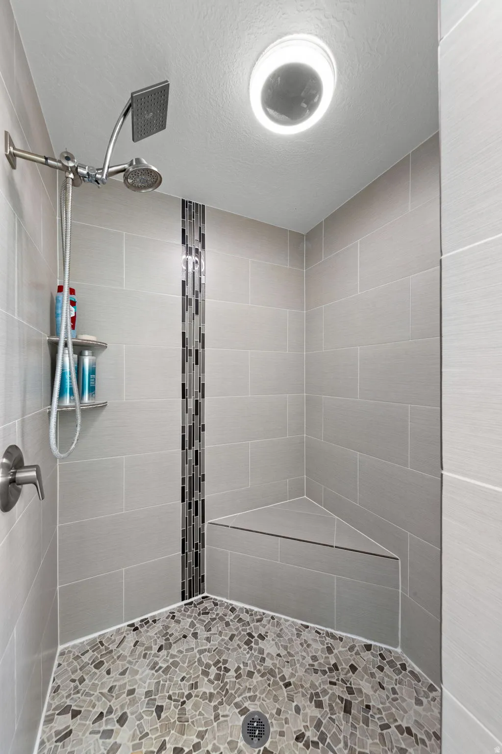 Walk-in shower featuring gray tile walls with a mosaic accent strip, a built-in corner bench, a pebble-finish floor, and a dual shower head system