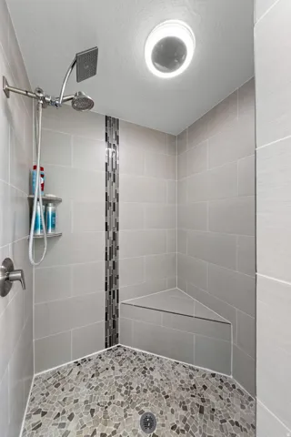 Walk-in shower featuring gray tile walls with a mosaic accent strip, a built-in corner bench, a pebble-finish floor, and a dual shower head system