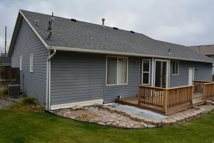 Rear exterior featuring gray horizontal siding, an attached wood deck with railings, sliding glass door access, and a manicured lawn