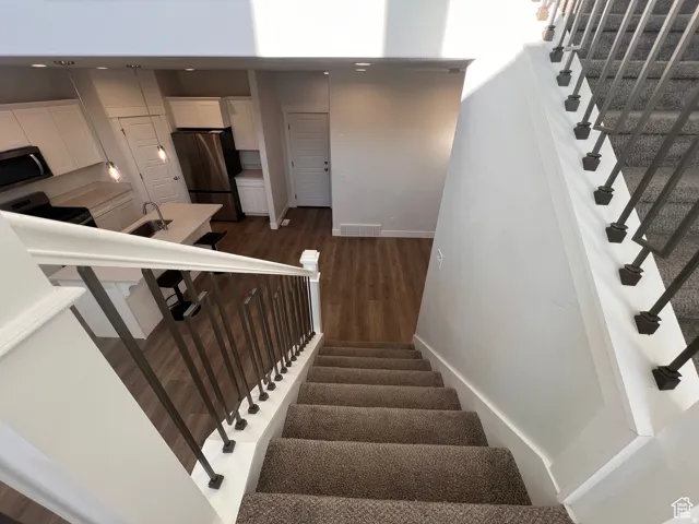 Carpeted staircase with dark metal balusters and white handrail leading down to a contemporary kitchen featuring white cabinetry, stainless steel appliances, a center island with sink, and wood-finish flooring