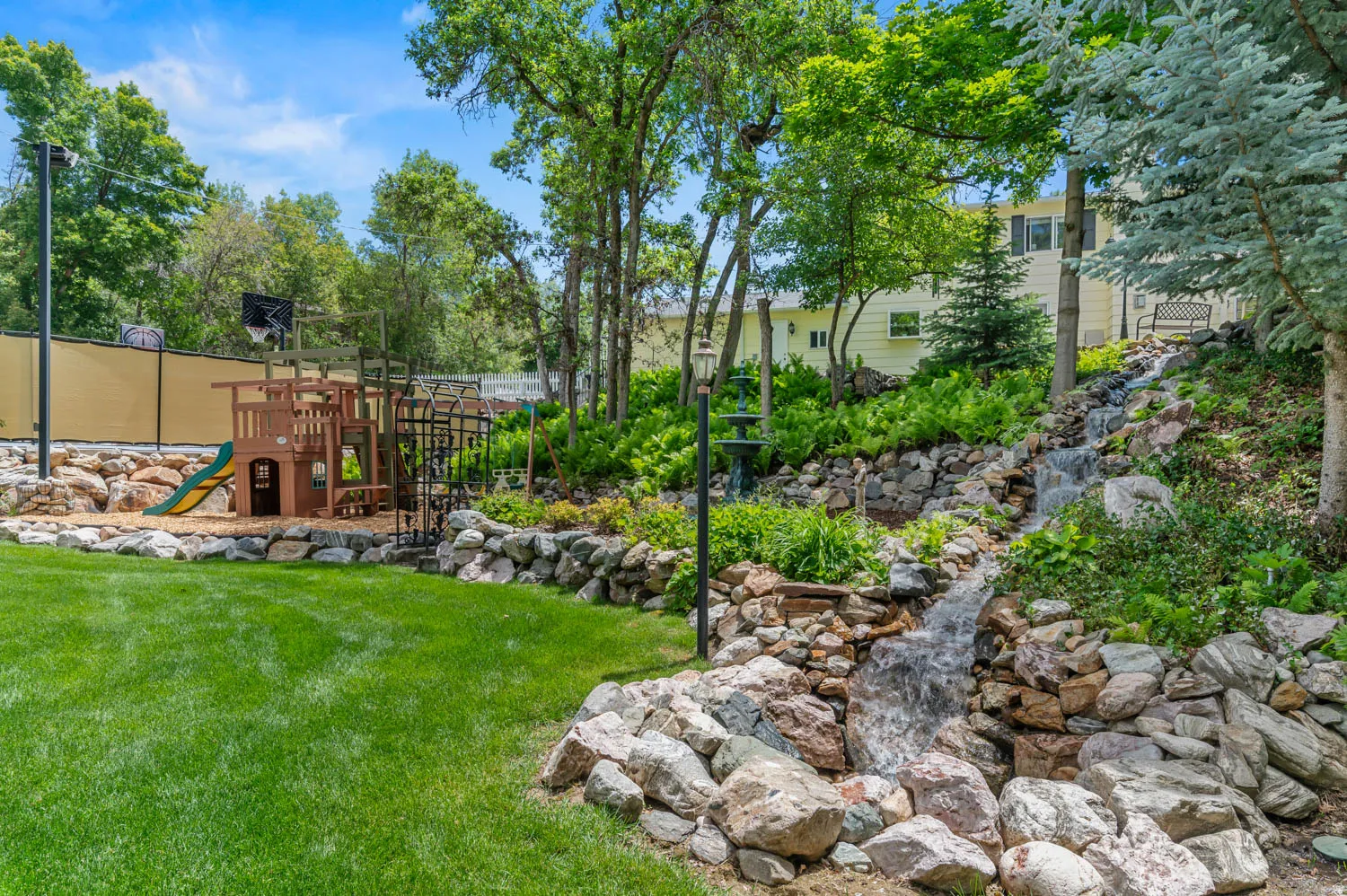 Expansive backyard featuring a multi-tiered rock waterfall, lush green lawn, and mature trees