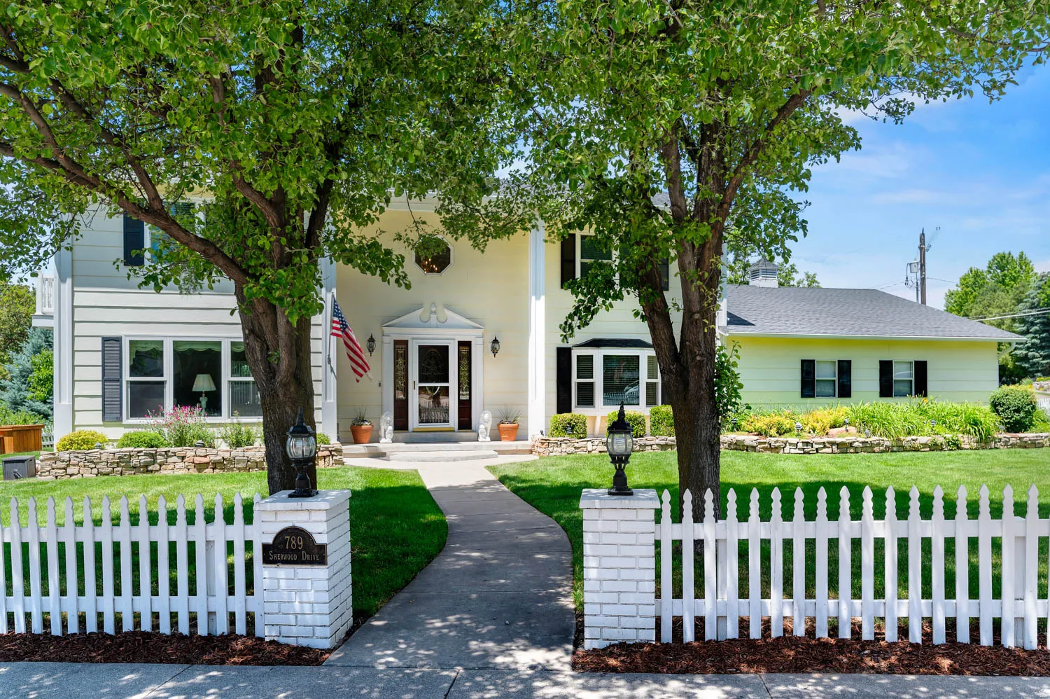 Two-story residence featuring a classic picket fence, a manicured lawn, mature trees, and a stone-lined garden