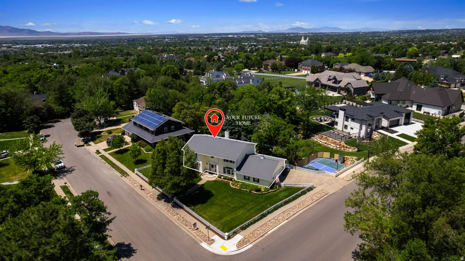Corner lot residence featuring a light-colored exterior, green lawn, mature trees, and a paved driveway