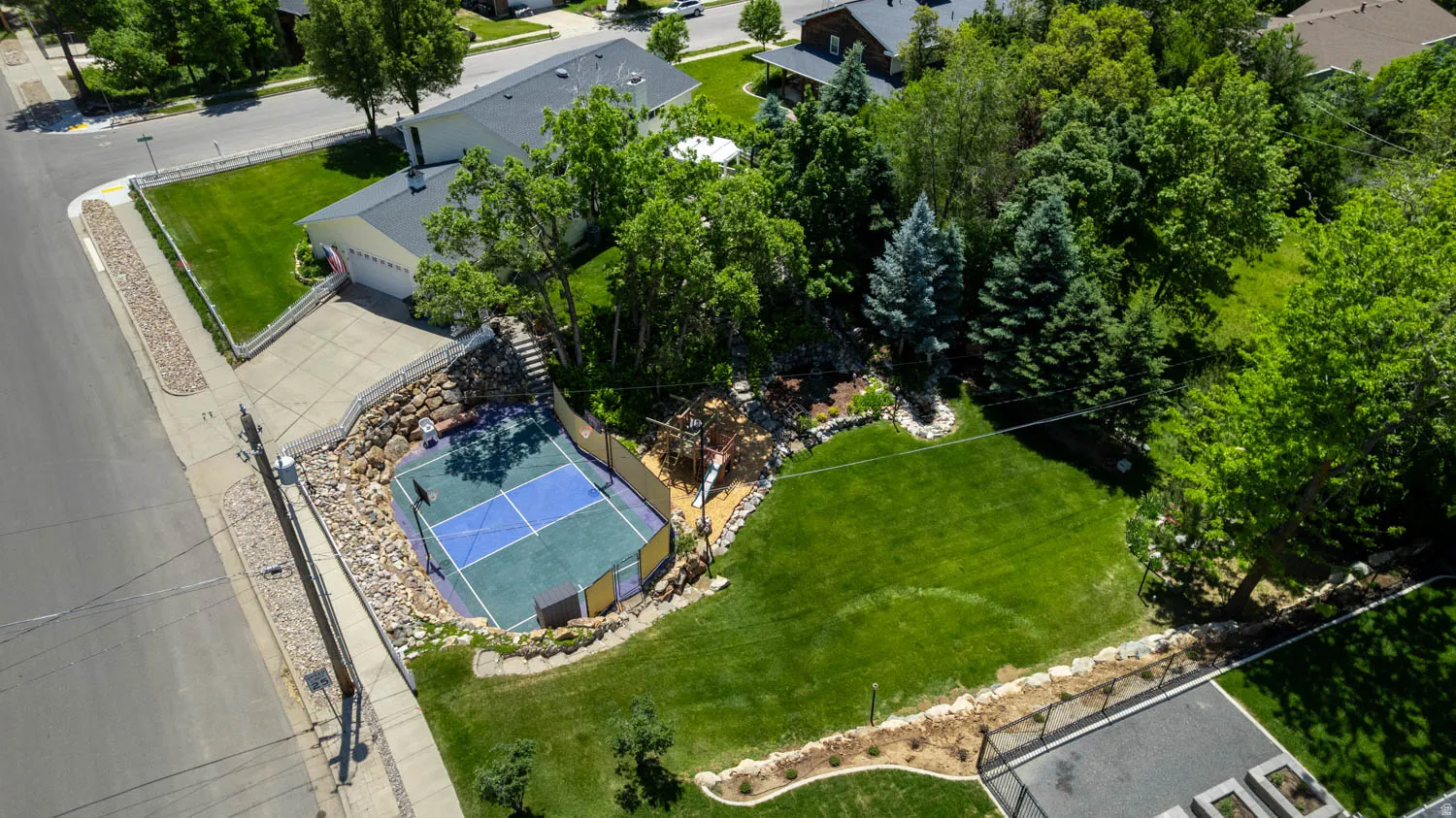 Expansive outdoor recreation area featuring a multi-sport court, established landscaping, mature trees, and a spacious lawn