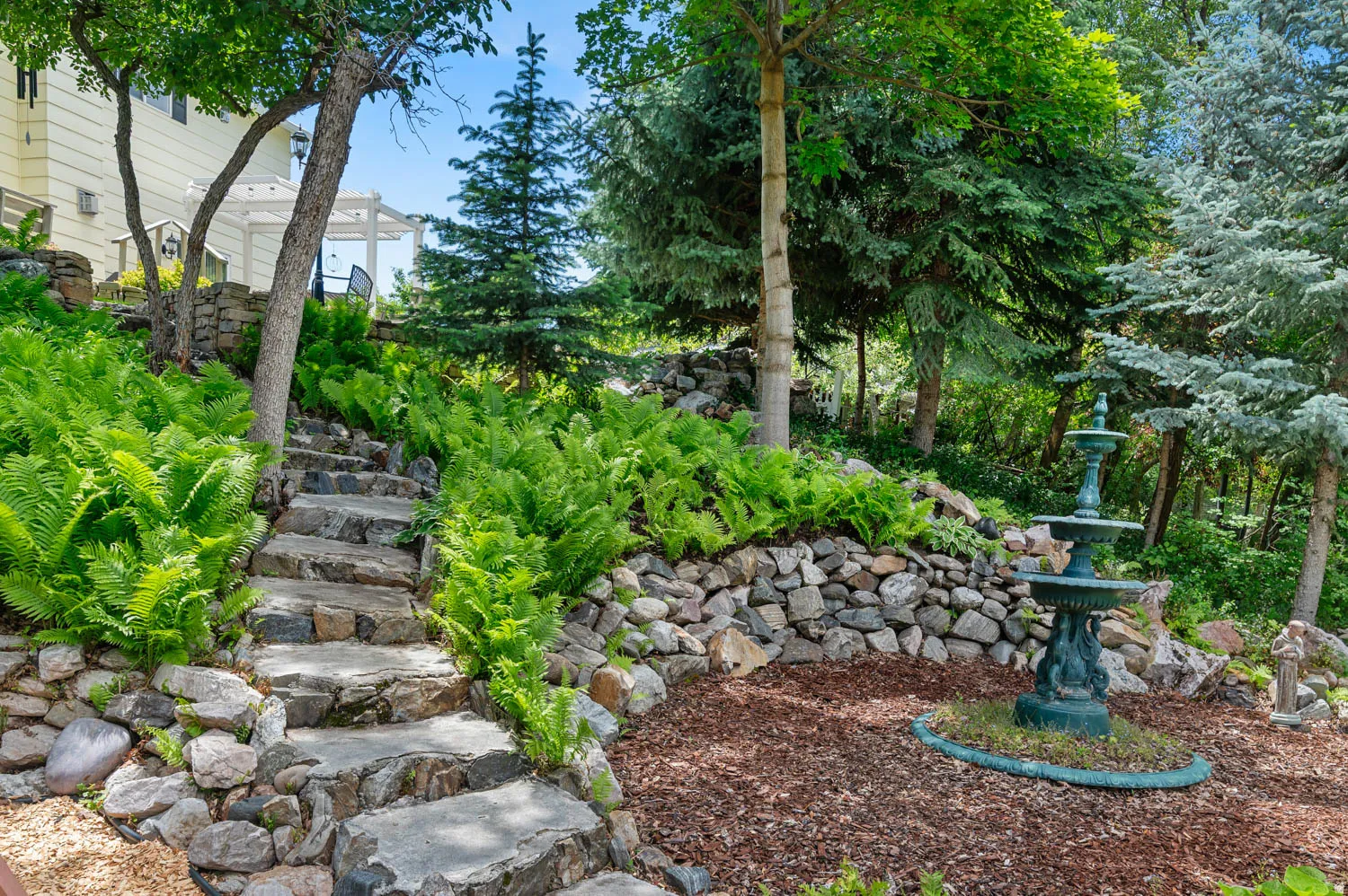 Landscaped garden featuring stone steps, mature trees, lush ferns, a decorative fountain, and a white pergola in the background