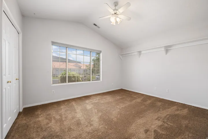 Vaulted ceiling room featuring a large multi-pane window, ceiling fan with integrated lighting, built-in shelving, plush brown carpet, and bifold closet doors