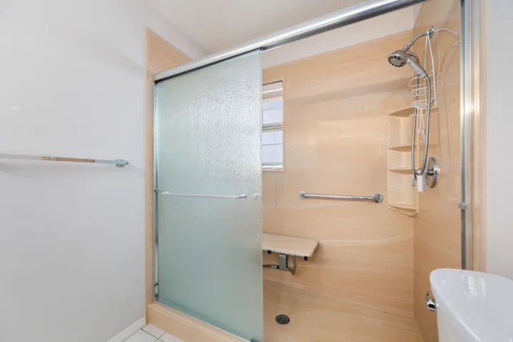 Walk-in shower featuring a sliding frosted glass door, light-toned shower surround, built-in shelving, chrome fixtures, and a folding shower bench