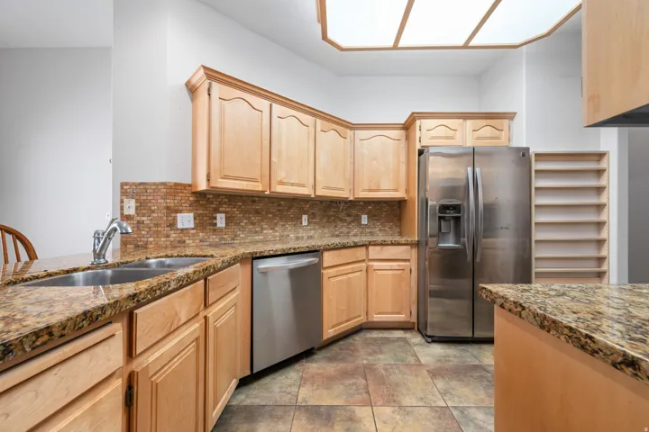 Well-appointed kitchen featuring light wood cabinetry, stone-finish countertops, stainless steel appliances, a mosaic tile backsplash, and a built-in pantry shelving unit