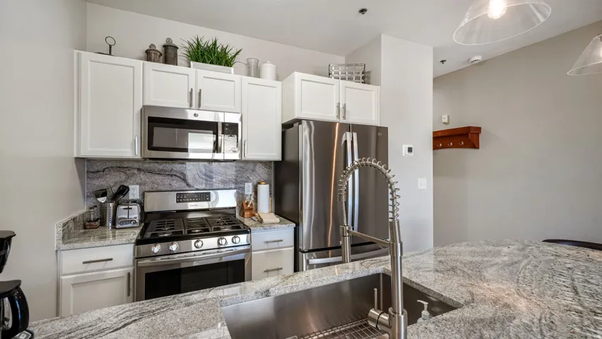 Kitchen featuring white cabinetry, stainless steel appliances, a deep stainless steel sink with a high-arc faucet, and light-toned countertops with dark veining