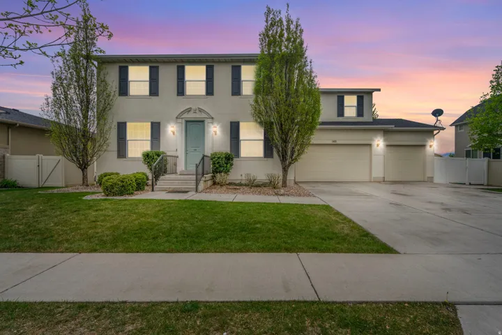 Two-story stucco residence featuring a light blue front door, black shutters, two prominent front trees, a manicured lawn, and a multi-car garage with a concrete driveway