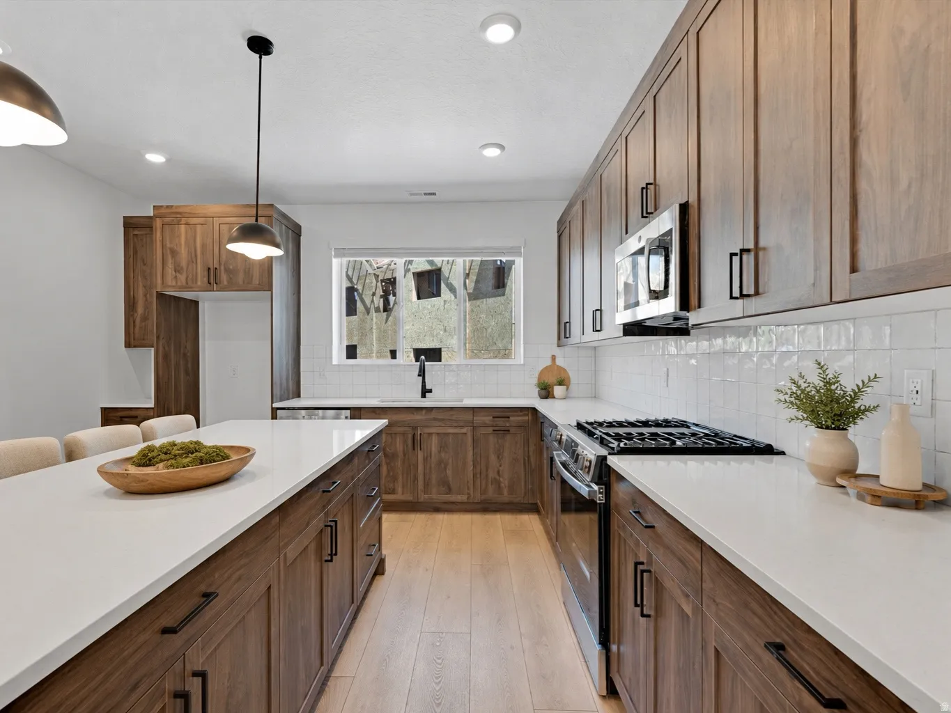 Modern kitchen with an elongated island, light-toned countertops, wood-finish cabinetry, a white subway tile backsplash, and stainless steel appliances