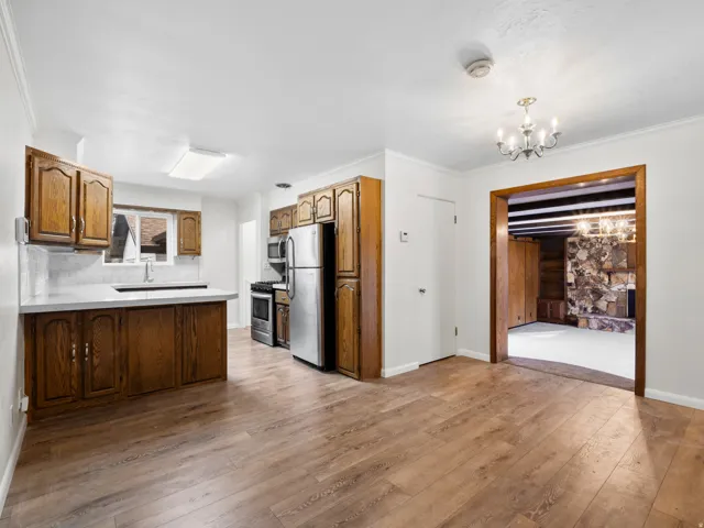 Open-concept living space featuring wood-finish flooring, a kitchen with wood cabinetry, stainless steel appliances, and a light-toned countertop, and an entryway to a room with a stone-clad fireplace