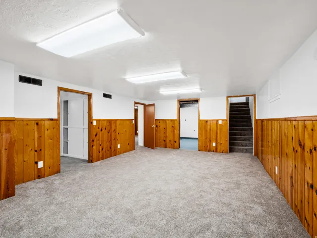 Spacious room featuring light gray carpet, natural wood paneling, white walls, and multiple recessed fluorescent light fixtures