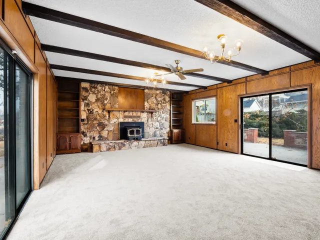 Spacious room featuring a stone fireplace, wood paneling, exposed ceiling beams, built-in shelving, and dual sliding glass doors