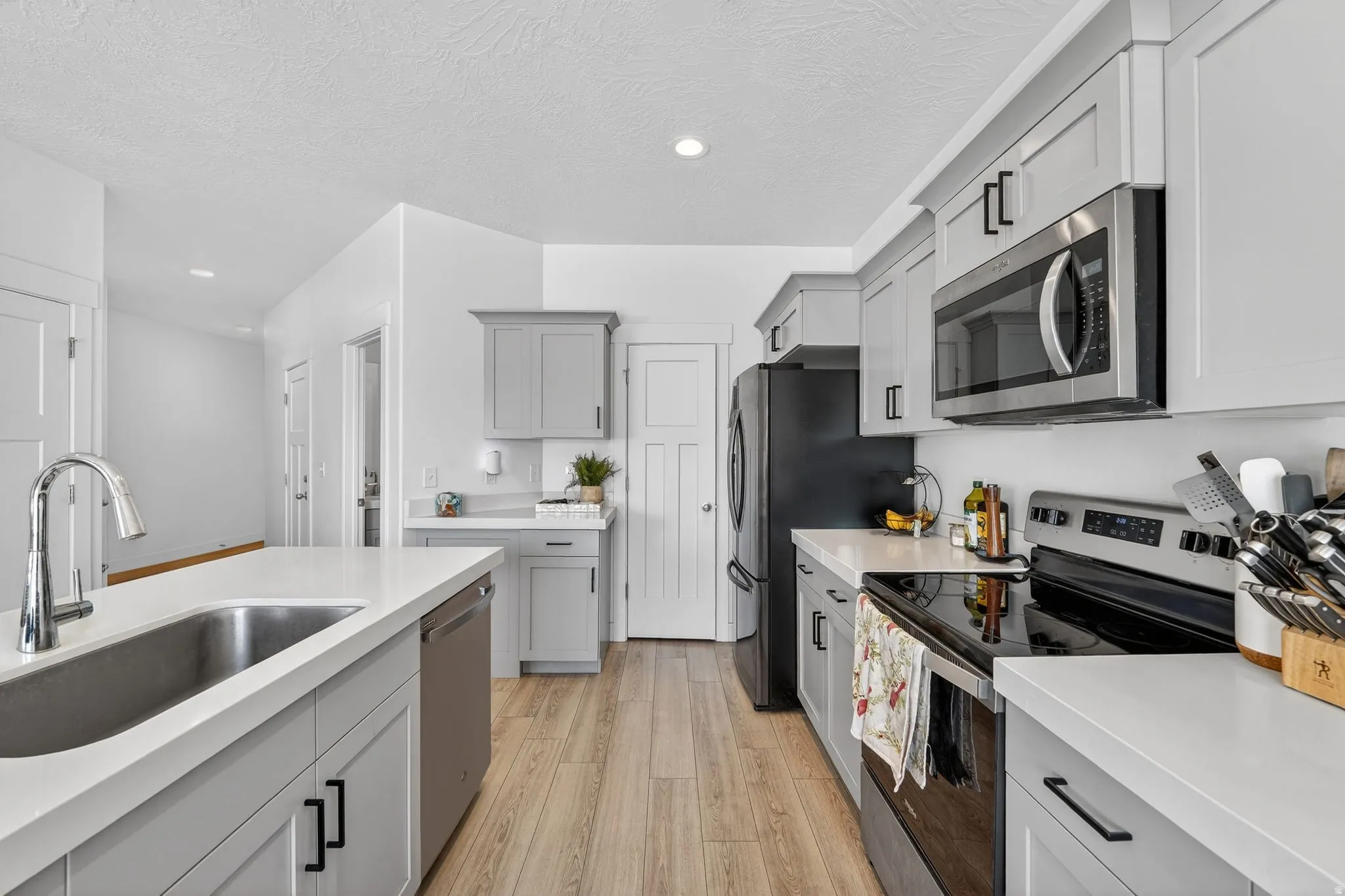 Kitchen featuring light grey cabinetry with black hardware, white countertops, wood-finish flooring, a stainless steel sink with a pull-down faucet, and built-in stainless steel appliances including a microwave and dishwasher