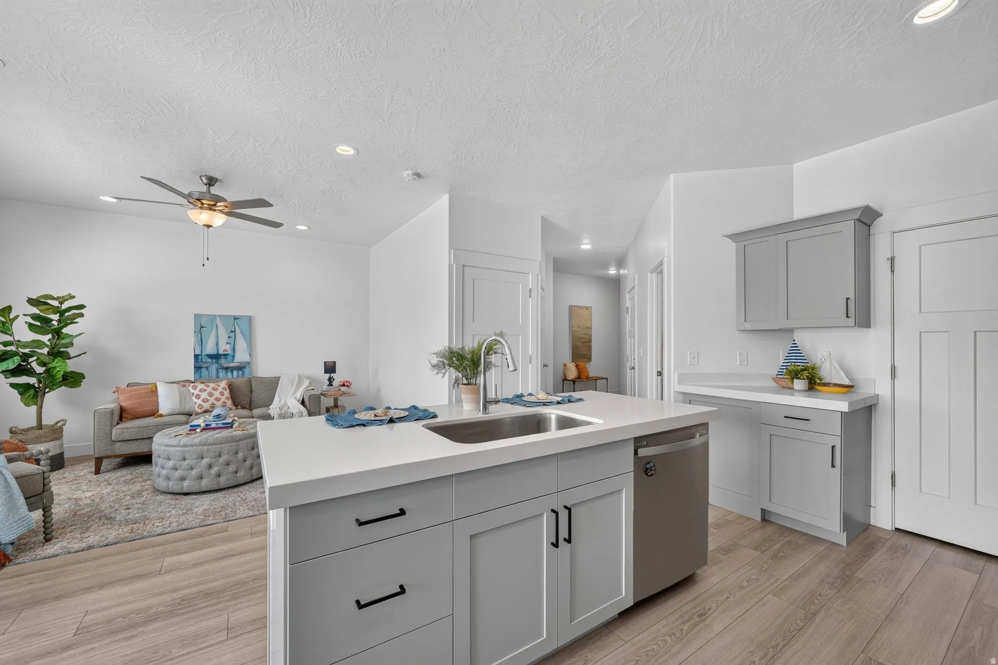 Open-concept living space featuring a central island with an undermount sink, light gray cabinetry, stainless steel dishwasher, and wood-finish flooring