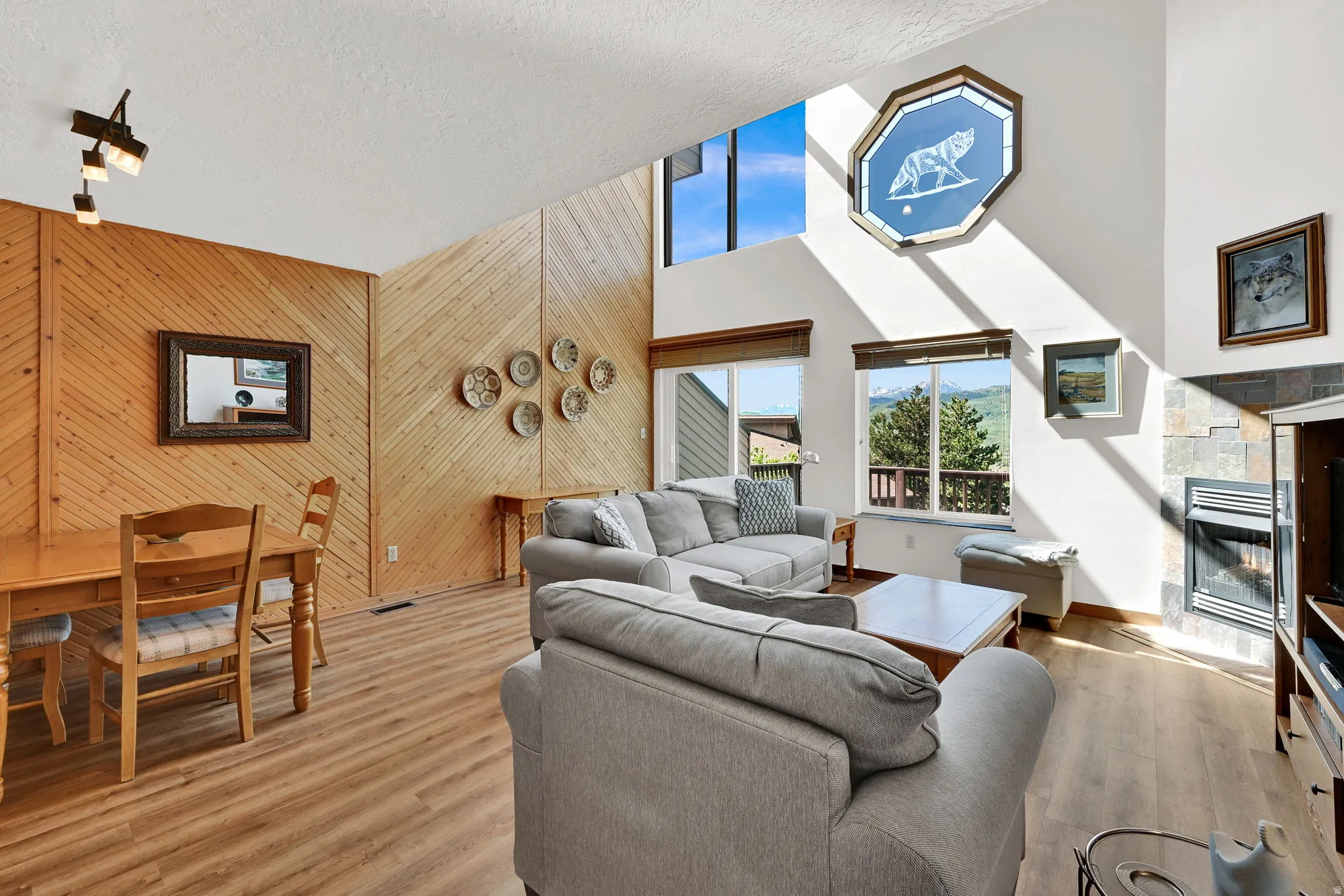 Vaulted ceiling living space featuring wood-finish flooring, a stone-clad fireplace, and multiple windows showcasing mountain scenery
