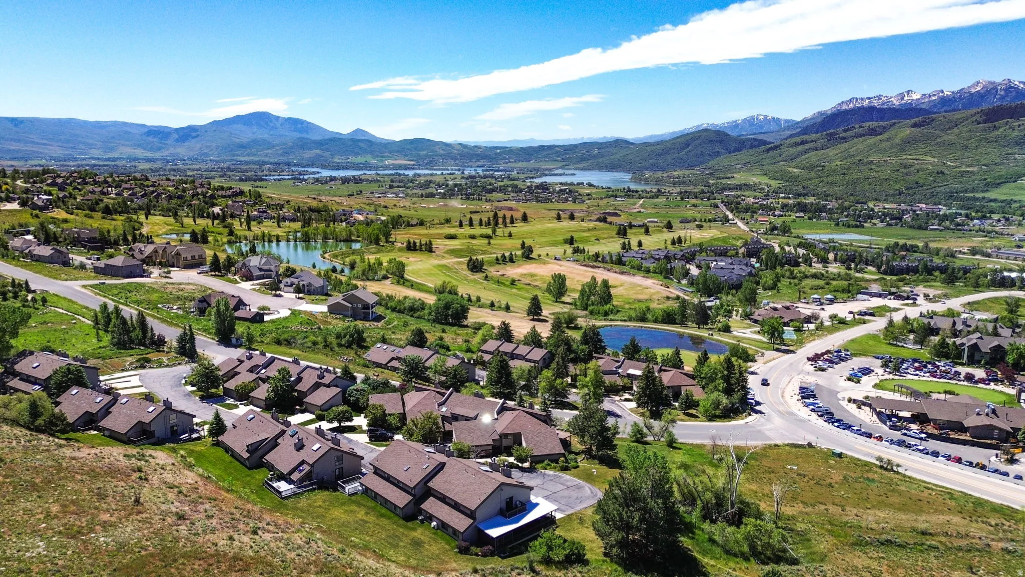 Expansive aerial view showcasing residential properties, a golf course with water features, distant lakes, and snow-capped mountains under a clear sky
