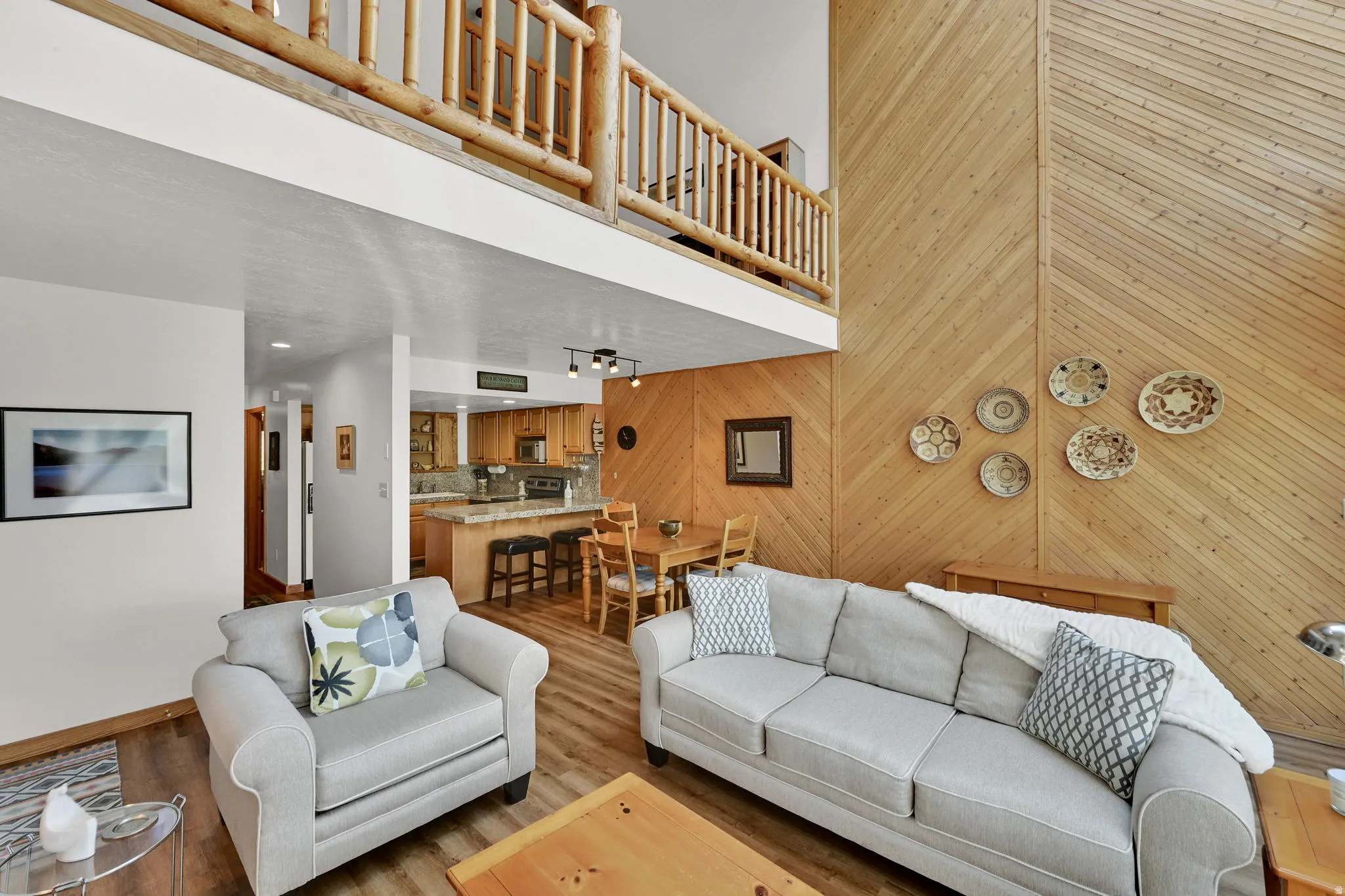 Living area featuring wood-finish flooring, a wood-paneled accent wall, a built-in kitchen island with seating, an open-concept layout, and a lofted space with a log railing
