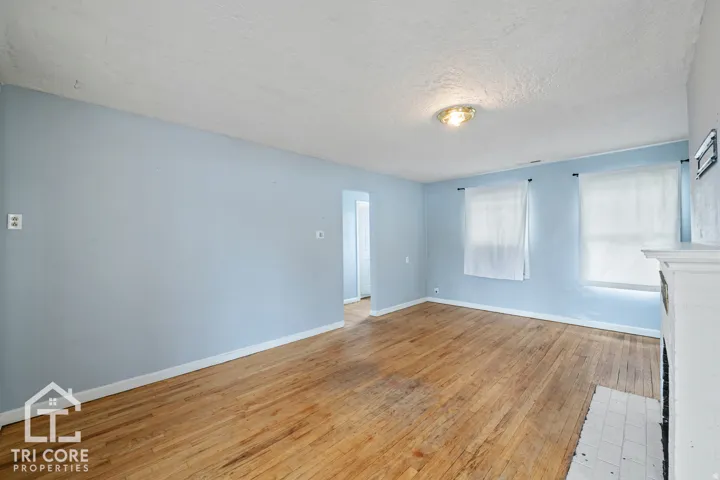 Spacious room featuring wood-finish flooring, two windows, a white baseboard trim, a central ceiling light fixture, and a fireplace with a white mantel and hearth