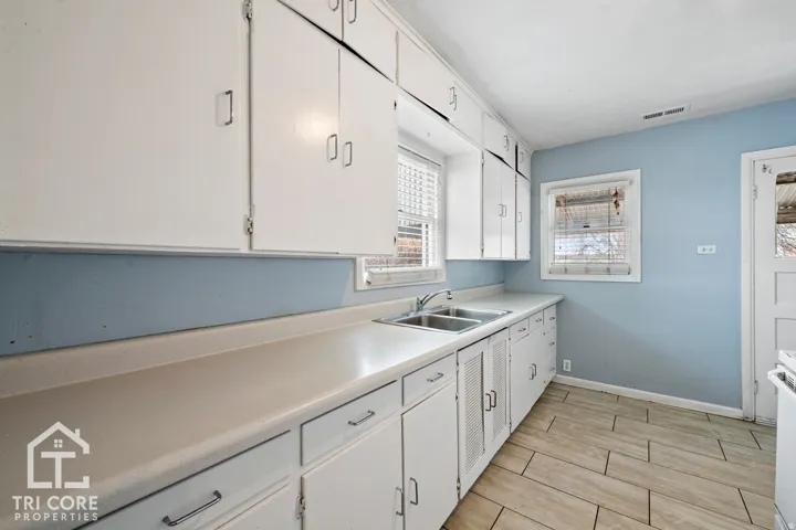 Kitchen featuring extensive white cabinetry, a double basin stainless steel sink, light-toned countertops, and wood-finish tile flooring