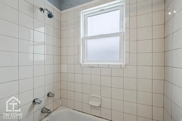Tub and shower combination featuring white square tiling, a chrome shower head, chrome faucet fixtures, and a window with privacy glass