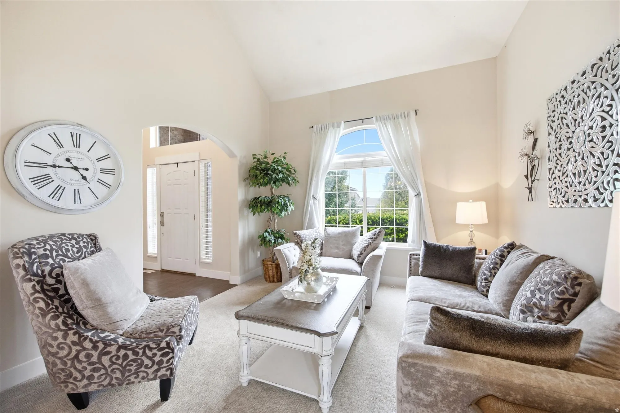 Living area featuring vaulted ceilings, an arched entryway, a large arched window with exterior landscaping, and plush carpet flooring
