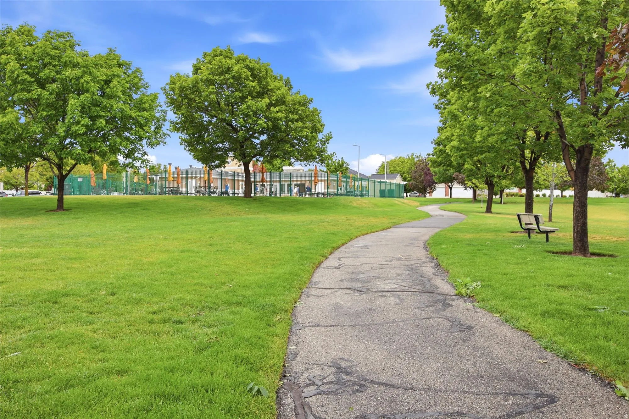 Green space featuring a winding paved pathway, mature trees, and a distant playground area