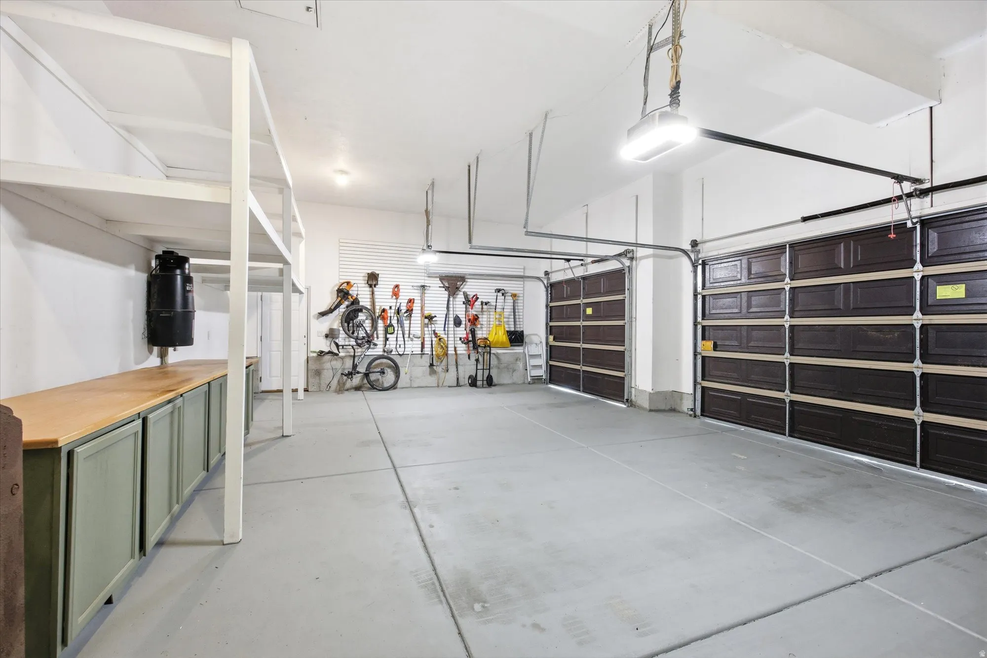 Spacious garage featuring two dark wood-paneled overhead doors, a light gray concrete floor, built-in shelving, and extensive cabinetry with wood countertops