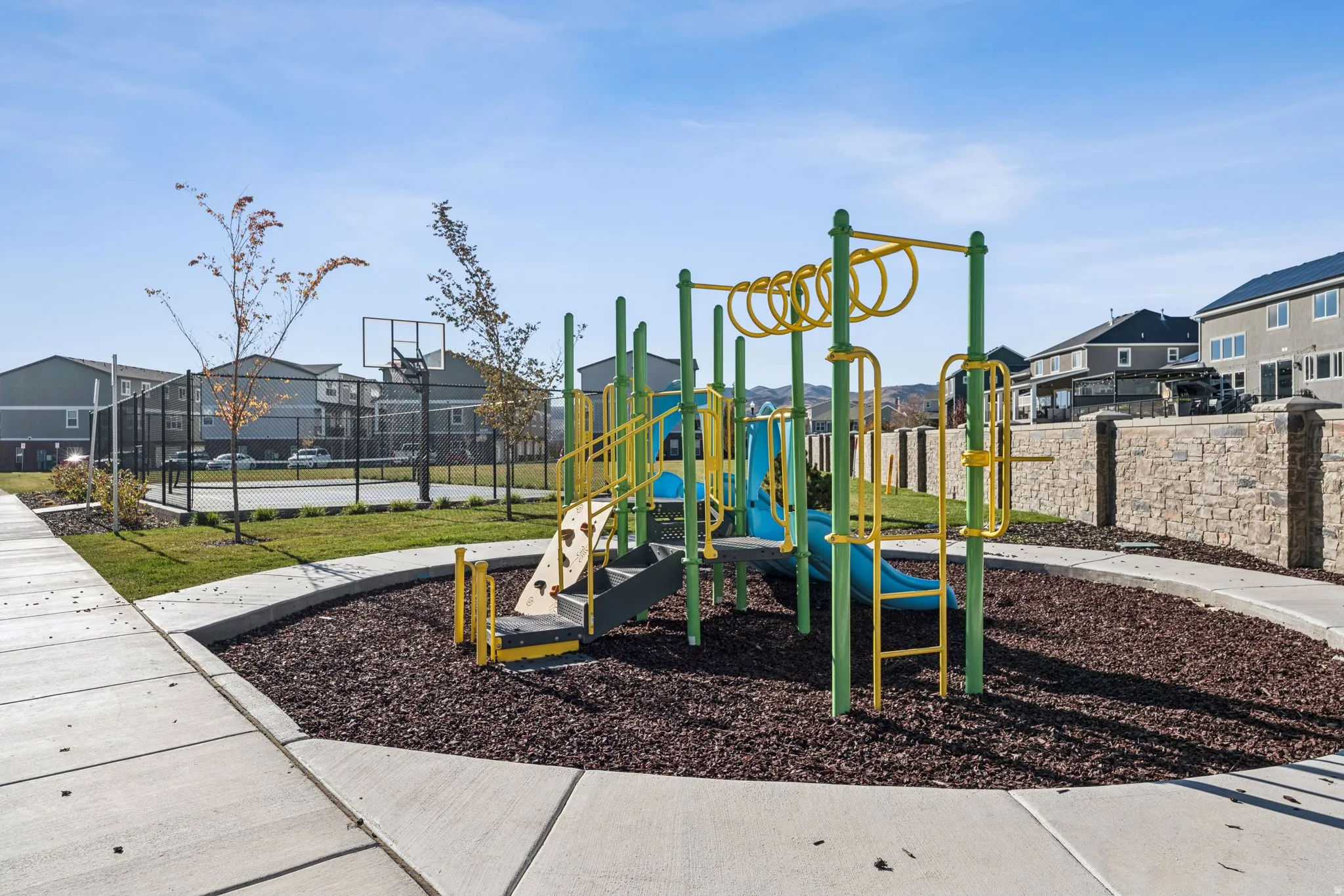 Recreational area featuring a playground with slides and climbing elements, a basketball court with a chain-link fence surround, concrete pathways, manicured grass areas, and a stone retaining wall