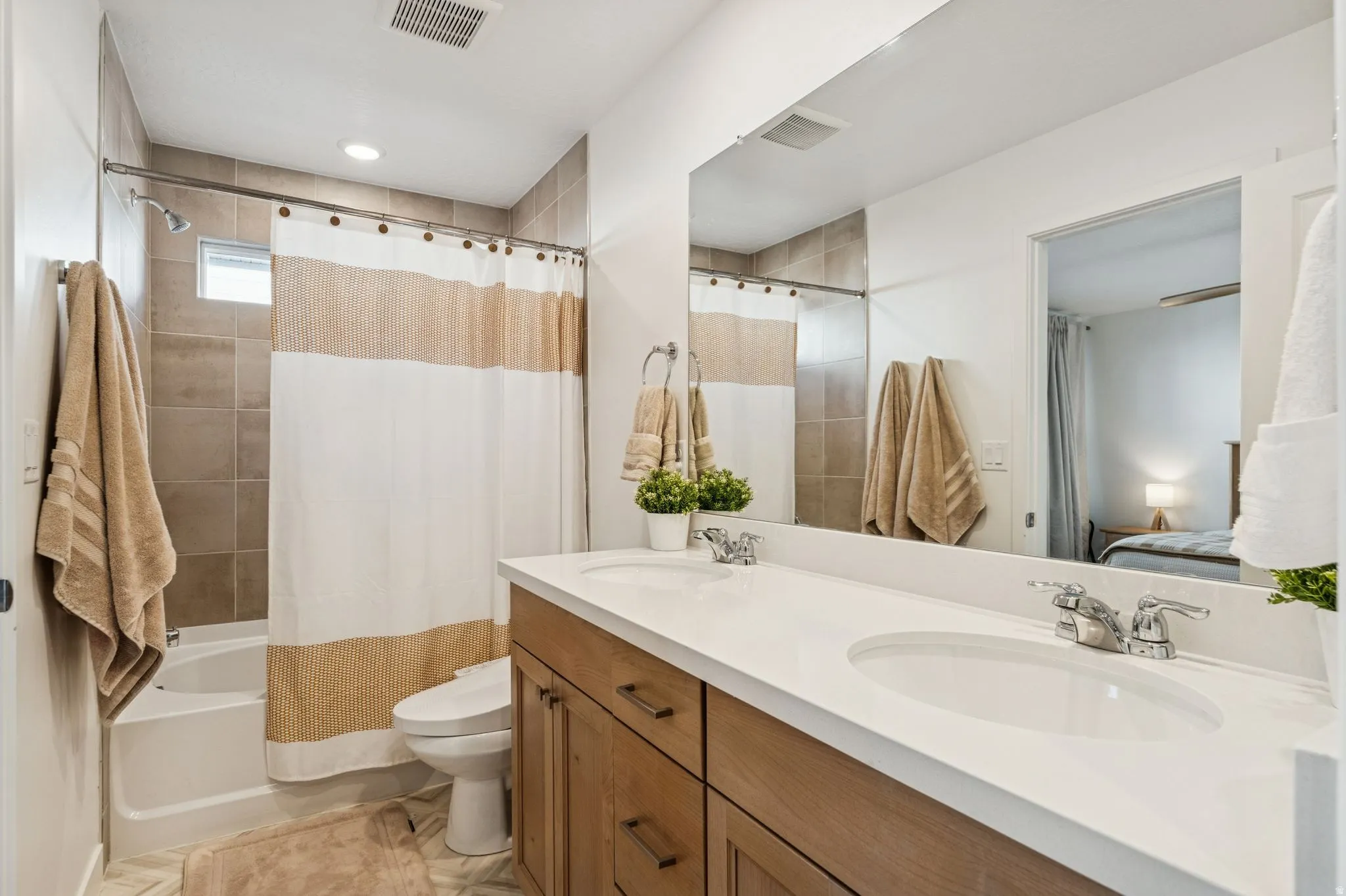 Bathroom featuring a dual-sink vanity with a white countertop and wood-finish cabinetry, a large frameless mirror, and a tiled tub/shower combination with a small window