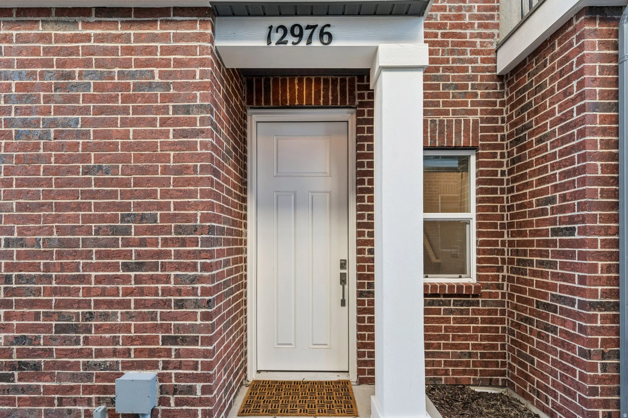 Brick exterior featuring a white paneled entry door with black hardware, a contrasting white column, and an overhead portico with address numbers
